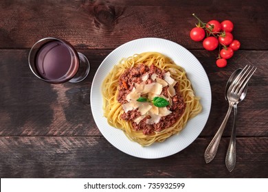 Overhead Photo Of Plate Pf Bolognese Pasta With Grated Parmesan Cheese And Fresh Basil Leaves, With Fork And Spoon, Cherry Tomatoes And Glass Of Red Wine, On Dark Rustic Texture With Copy Space
