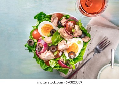 Overhead Photo Of A Plate Of Salad With Canned Tuna, Boiled Eggs, Green Lettuce Leaves, Onions, Black Olives, And Cherry Tomatoes, On Teal Texture With Glass Of Rose Wine, White Sauce, And Copy Space