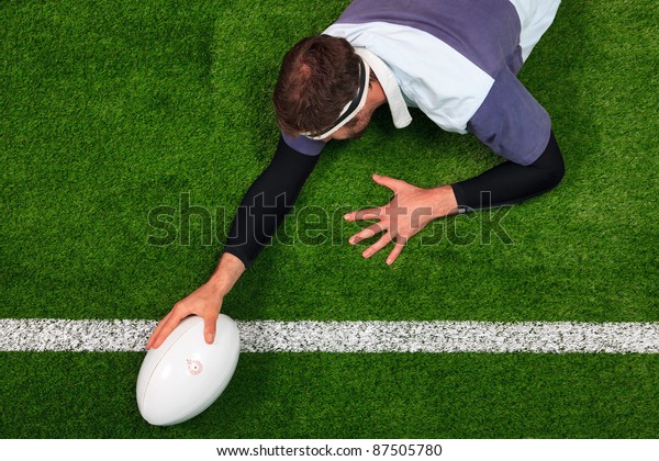 Overhead photo of a rugby\
player stretching over the line to score a try with one hand on the\
ball.