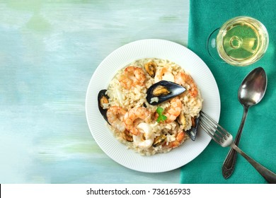 An Overhead Photo Of A Seafood Risotto On Teal Textures, With A Fork And A Spoon, A Glass Of White Wine, And A Place For Text