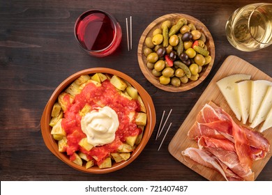 An Overhead Photo Of Traditional Spanish Tapas. Pickled Olives, Patatas Bravas, Potatoes With A Red And White Sauce, Jamon, And Cheese, With Glasses Of Wine, Shot From Above On A Dark Rustic Texture