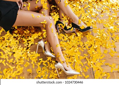 Overhead portrait of girls wearing elegant high heel shoes and sitting on the floor during party. Ladies posing on the sparkle golden confetti, lying on the ground.