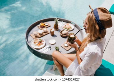 Overhead Portrait Of Smiling Blonde Woman In Brown Hat Drinking Cocktail In Pool. Outdoor Shot Of Excited Female Model Chilling At Resort With Fruits.