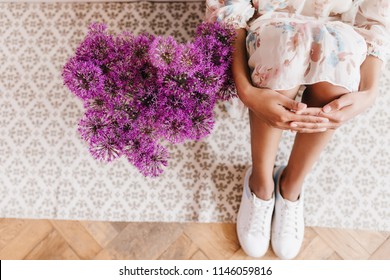 Overhead Portrait Of Trendy Tanned Girl Sitting On The Floor With Purple Bouquet. Indoor Shot Of Relaxed Female Model In White Shoes And Dress Posing Beside Flowers.