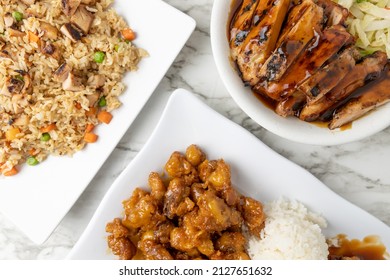 Overhead Shot Of Asian Food Dishes Including Teriyaki Chicken, Sweet And Sour Chicken, And Chicken Fried Rice On White Plates Indoors On A Marble Table Surface.