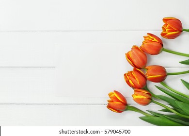 Overhead shot a bouquet of orange and yellow Spring tulip flowers and blank paper card for Mother's or Women's Day over white wood table top. Flat lay top view style.