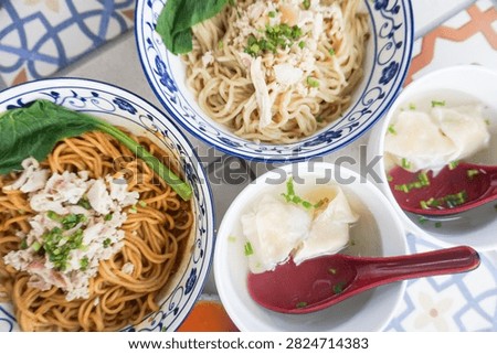 An overhead shot captures two large bowls of Asian noodles, one with a light broth and the other with a rich, dark sauce, accompanied by two small bowls of steamed dumplings, creating a delightful an.