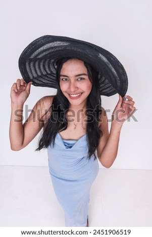 Overhead shot - A cheerful young woman poses in a stylish baby blue dress and oversized black straw hat, isolated against a white backdrop.