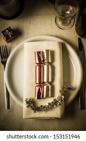 Overhead Shot Of A Christmas Table Setting With Shiny Foil Crackers And Starry Tinsel On A Dinner Plate With A Small Gift Box For The Diner.  A, Wine Glass And Bottle And A Candle Sit At Top Of Frame.