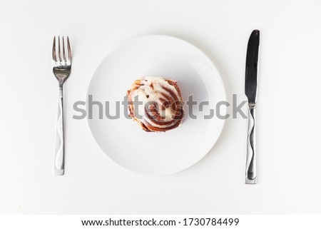 Overhead shot of cinnabon roll or cinnamon, homemade sweet traditional dessert bun with white cream sauce on white plate on the white background with 
cutlery