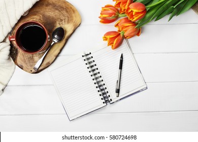 Overhead Shot A Cup Of Coffee, Open Book, Knit Throw Blanket And A Bouquet Of Orange And Yellow Tulip Flowers Over White Wood Table Top. Flat Lay Top View Style.