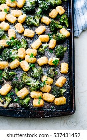 Overhead Shot Of Dark Green, Rich Broccoli And Tater Tots On Black Metal Roasting Pan Topped With Chopped Parsley And Fresh Parmesan Cheese With Bright Lighting On Cream Marble Surface With Blue Linen