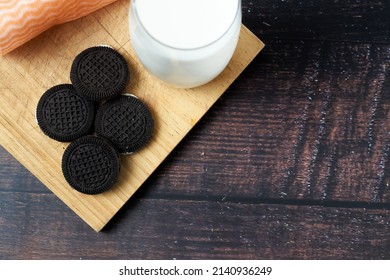 Overhead Shot Of Delicious Chocolate Cookies With Vanilla Cream And A Glass Of Milk On A Wooden Board And Dark Background