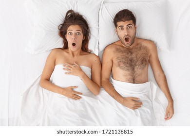 Overhead Shot Of Emotional Shocked Woman And Man Stay In Bed Covered By Sheet, Stare With Wide Opened Mouth, Overslept Work Or Flight. Shocked Family Couple Awake In Hotel Room. Astounded Lovers