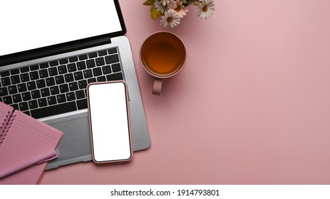 Overhead shot of feminine workspace with smart phone, laptop, notebook and copy space on pink background.