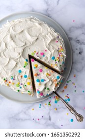 Overhead Shot Of Of A Festive White Birthday Cake With One Slice Cut Out. A Vintage Serving Utensil Is Pulling Out A Slice With Sprinkles Scattered Around.