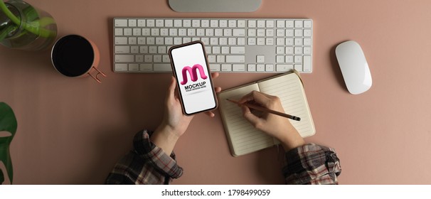 Overhead shot of freelancer using mock up smartphone and taking note while working on computer desk 