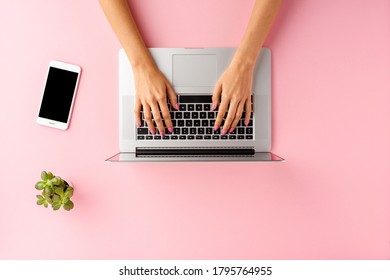 Overhead Shot Of Woman’s Hands Working On Laptop On Pink Table With Mobile Phone And Flower. Office Desktop. Flat Lay