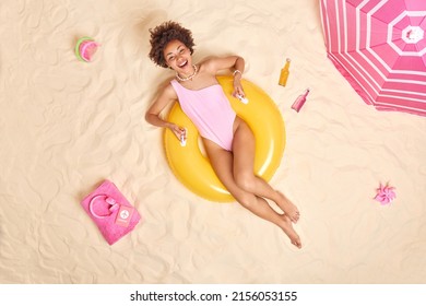 Overhead Shot Of Happy Curly Haired Woman In Swimwear Poses On Yellow Inflated Swimring Spends Free Time At Beach Lies In Sun Surrounded By Sand Toys Bottle Of Energetic Drinks Headphones On Towel