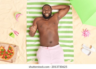 Overhead Shot Of Happy Handsome Man With Dark Skin Clenches Teeth And Fist Reacts On Successful Deal Poses On Striped Towel At Sandy Beach Spends Summer Vacation At Seaside. Nice Vacation Concept