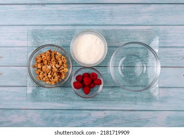 Overhead Shot Of Ingredients For A Healthy Breakfast On A Glass Tray. Including Cereals, Yogurt, Raspberries And An Empty Glass Bowl, Placed On A Blue Wooden Table.