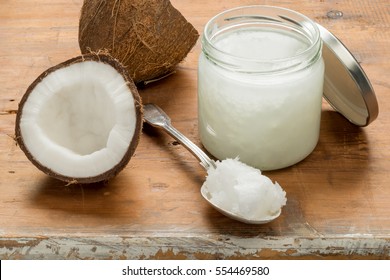 Overhead shot of a jar of coconut oil with a dessetspoon of oil  and a halved coconut to the left on a wooden background