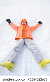 Overhead Shot Of A Little Girl Lying On Snow And Making A Snow Angel While Looking Up And Smiling At The Camera