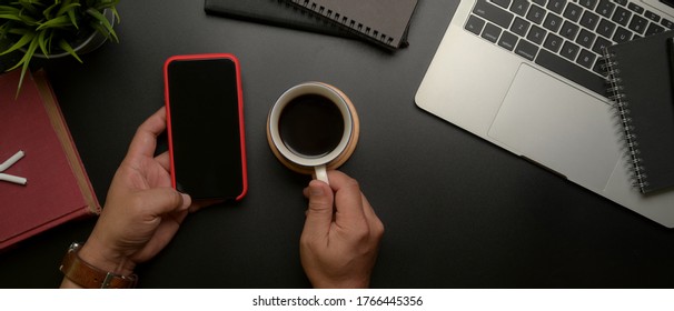 Overhead Shot Of Male Entrepreneur Left Hand Using Smartphone While Right Hand Holding Coffee Cup On Dark Modern Office Desk