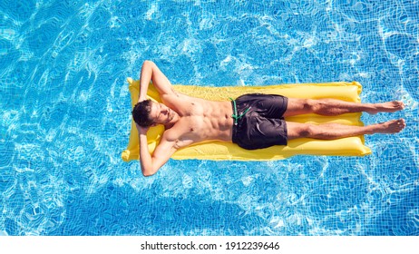 Overhead Shot Of Man In Swim Shorts Floating On Air Bed On Summer Vacation In Outdoor Swimming Pool