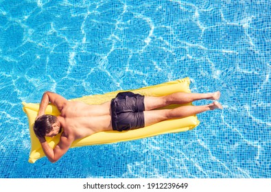 Overhead Shot Of Man In Swim Shorts Floating On Air Bed On Summer Vacation In Outdoor Swimming Pool