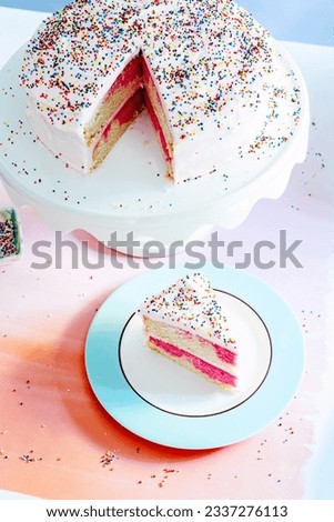 An overhead shot of an ombre vanilla and pink birthday cake with slice cut out on white cake stand with multicolored sprinkles, blue background and pink surface.