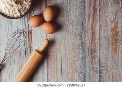 Overhead Shot On Weathered Wooden Work Table With Eggs, Whisk, Rolling Pin, Flour. Copy Space On The Right