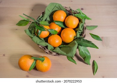 Overhead Shot On A Wicker Basket Full Of Fresh Clementines With Leaves, On Wooden Surface And Two Clementines Out Of The Basket