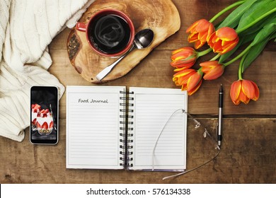 Overhead Shot Of An Open Food Journal Book With Cell Phone, Coffee And Flowers Over A Wood Table Top Ready To Plan Diet. Flat Lay Top View Style.