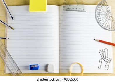 Overhead Shot Of An Opened School Exercise Book. Surrounded By Stationery Items On A Light Wood Desk. Lined Pages Provide Copy Space.