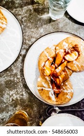 Overhead Shot Of Pan-fried Peaches On Sopapillas With Goat's Milk Yogurt Drizzle Served On White Plates And Golden Beer On Dappled Gray Restaurant Table.