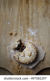 Overhead Shot Of A Partially Eaten Christmas Mince Pie With Crumbling Pastry And Dusting Of Icing Sugar, On An Old Wooden Chopping Board.