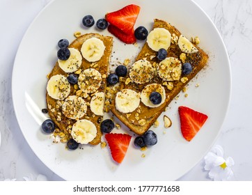 Overhead Shot Of Peanut Butter Banana Toast Topped With Seeds, Granola, And Berries On A White Plate On Marble