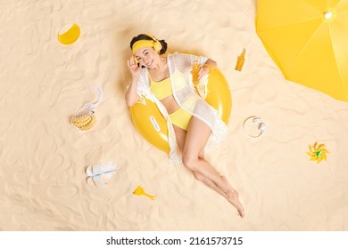 Overhead Shot Of Positive Attractive Asian Woman Dressed In Yellow Swim Suit Enjoys Listening Music Poses On Inflated Swimring Drinks Energetic Drink Chills At Sandy Beach Has Summer Vacation.