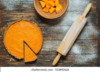 Overhead Shot Of Pumpkin Pie On Rustic Brown Table. Rolling Pin And Pumpkin Pieces In Clay Bowl On A Side. Spilled Flour. Pie Slice. Healthy Food, Baking, Cooking In Fall, Harvest, Thanksgiving.