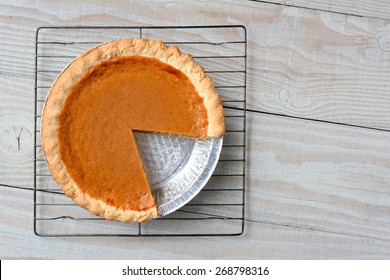 Overhead Shot Of A Pumpkin Pie With A Slice Cut Out On A Cooling Rack. Horizontal Format On A Rustic White Kitchen Table.