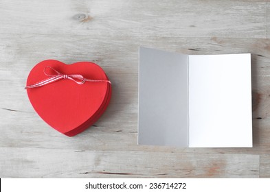 Overhead shot of a red heart-shaped gift box with ribbon bow, and a blank, opened greeting card. Both set on a light woodplank table.  Processed to give a very light, whitened feel.