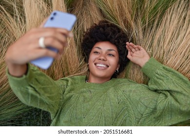 Overhead Shot Of Relaxed Afro American Woman Lies In Long Grass Enjoys Nature Holds Smartphone Takes Selfie Uses Front Mobile Camera Smiles Broadly Wears Casual Green Jumper Shoots Video For Blog