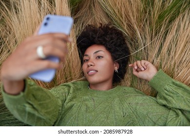 Overhead Shot Of Relaxed Curly Haired Young Woman Dressed In Casual Jumper Lies Relxed In Long Grass Holds Mobile Phone Makes Selfie Portrait Spends Free Time Outdoors. Millennial Content Maker