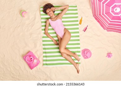 Overhead shot of relaxed young woman in bathingsuit lies in sun during sunny day surrounded by bottles of energetic drinks headphones sand toys spends summer vacation at seaside. Perfect holiday
