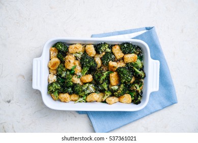 Overhead Shot Of Rich Green Broccoli And Roasted Tater Tots Casserole Topped With Grated Parmesan Cheese And Black Pepper On Light Blue Linen In Simple White Casserole Dish On Cream Marble Surface.