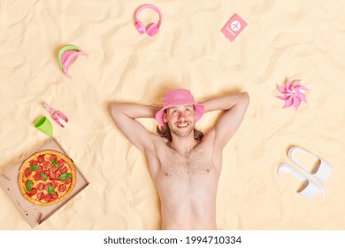 Overhead Shot Of Shirtless Bearded Redhead Man Wears Pink Panama Keeps Hands Behind Head Smiles Gladfully Lies On White Sand Surrounded By Beach Accessories Enjoys Leisure Day Summer Holidays.