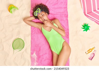 Overhead Shot Of Sleepy Female Model Wears Green Swim Suit And Snorkeling Mask On Forehed Yawns Covers Mouth With Hand Lies In Sun Poses At Beach Surrounded By Sand Toys Bottles Of Energetic Drinks