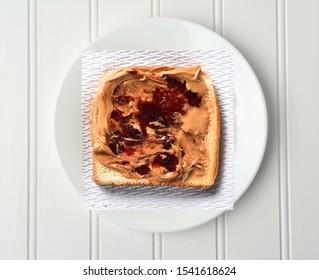 Overhead Shot Of A Slice Of Toast With Peanut Butter And Jelly, On A Paper Napkin And White Plate.