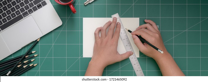 Overhead Shot Of Student Cutting Paper With Cutter On Cutting Mat With Laptop, Stationery And Coffee Cup